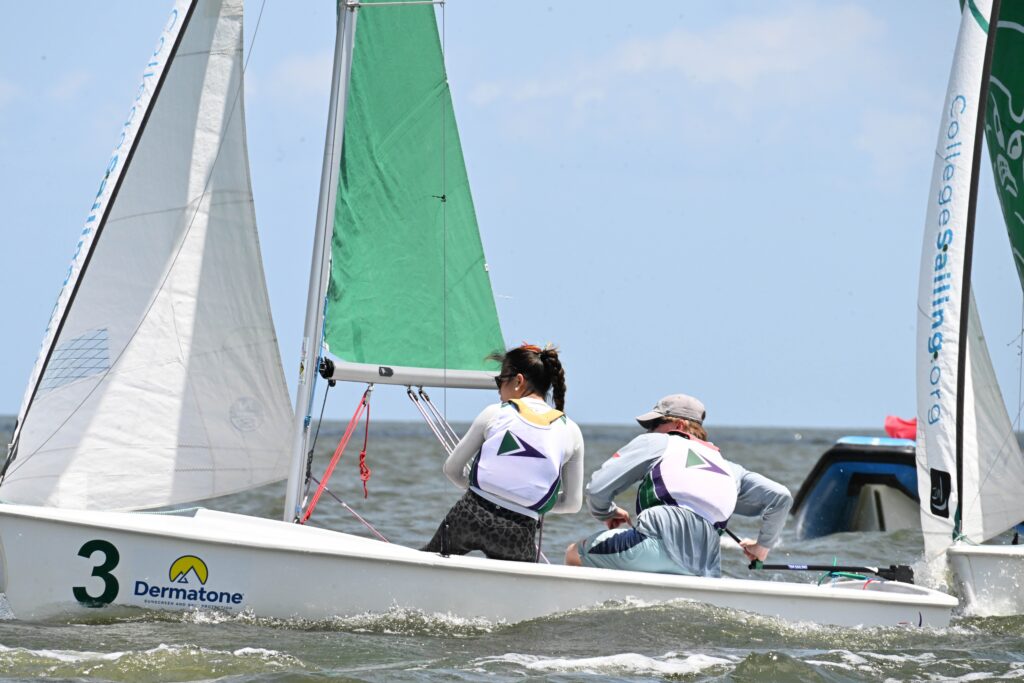 Two sailors wearing life vests race a small sailboat marked with the number 3 on the water. The boat has green and white sails, and there is another boat visible in the background under a blue sky.
