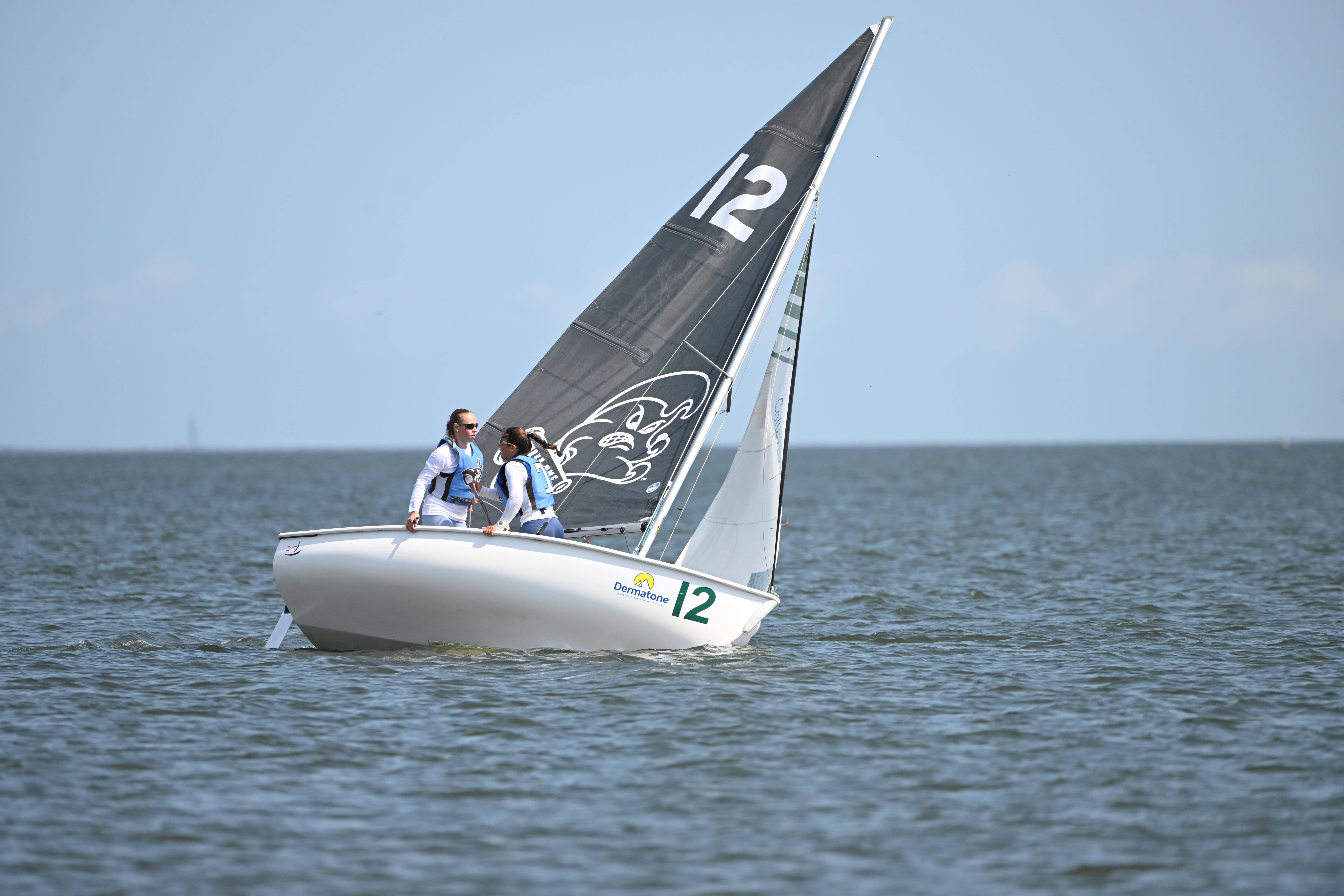 Two people wearing life jackets sail a white boat with a black sail marked “12” on calm water under a clear sky.