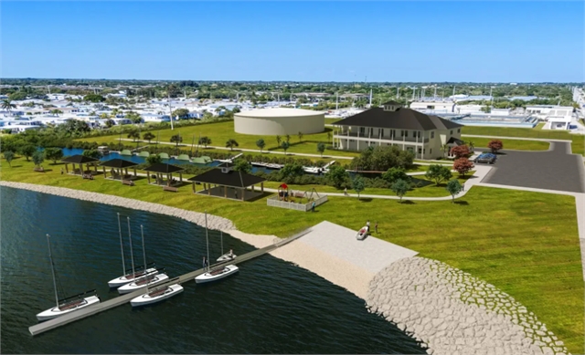 A waterfront facility with several small sailboats docked, green lawns, shaded pavilions, a large building, and a circular structure, with a cityscape visible in the background under a clear blue sky.