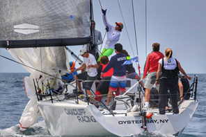 A group of sailors, wearing colorful clothing, actively maneuver a sailboat labeled "OYSTER BAY, NY" and "ONDARE RACING" during a race on open water under clear skies.