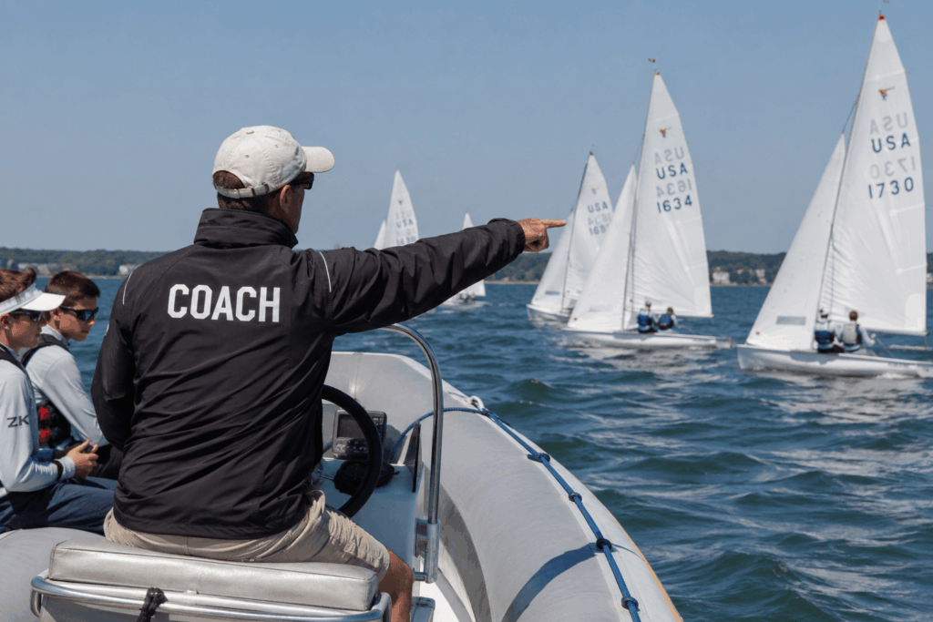 A sailing coach in a black jacket labeled 'COACH' sits on a motorboat, pointing toward sailboats racing on the water, with a crew of young sailors nearby under a clear blue sky.