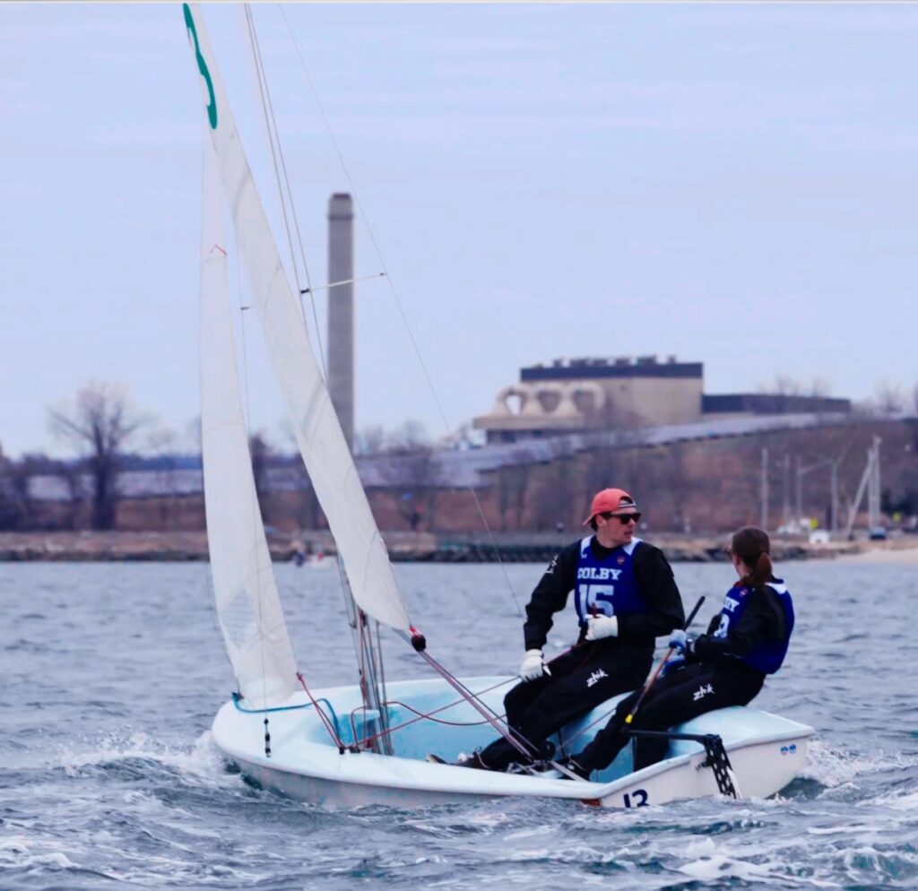 Two people in blue jerseys sail a small white boat numbered 13 on a body of water, with a shoreline, buildings, and leafless trees visible in the background under a cloudy sky.