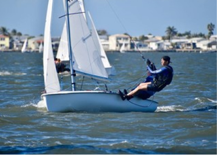 A person in a blue shirt and life jacket is sailing a small white sailboat on a sunny day, leaning out over the side as they steer. Houses and palm trees are visible onshore in the background.