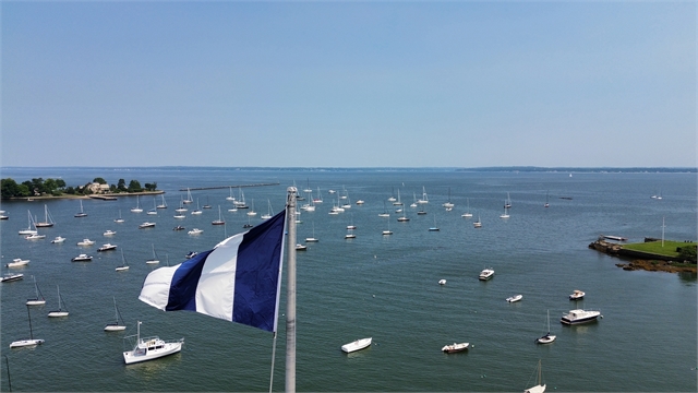 A blue and white flag waves in the foreground, overlooking a harbor filled with many anchored sailboats on calm water under a clear blue sky, with a shoreline and trees in the distance.