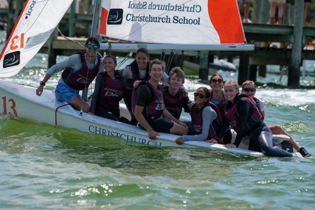 A group of smiling teenagers wearing life jackets sit together on a small sailboat labeled 'CHRISTCHURCH' on the water, with a dock and wooden pilings visible in the background.