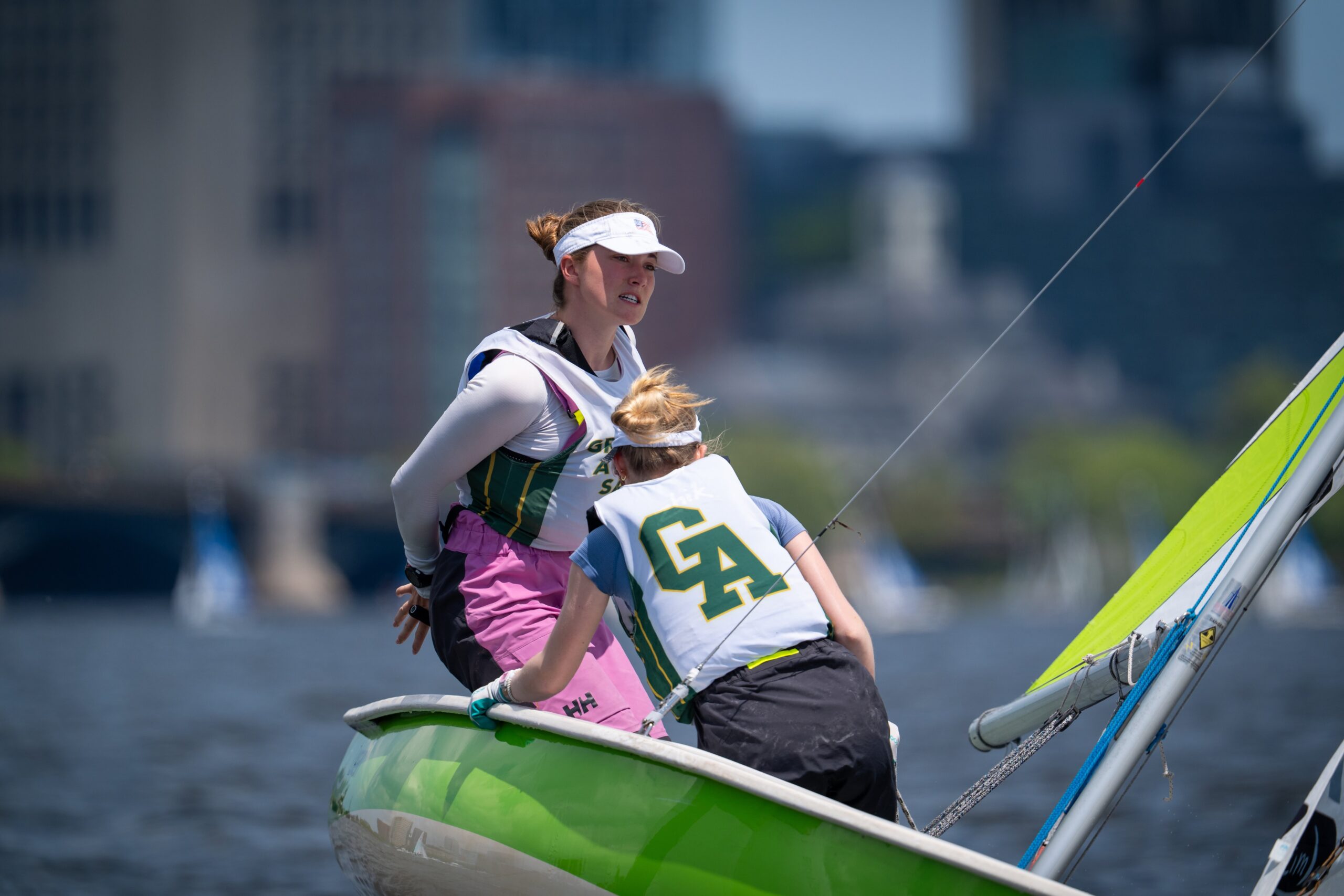 Two women sailing in a green and white boat, both wearing life vests and sports gear, with water and a cityscape blurred in the background on a sunny day.