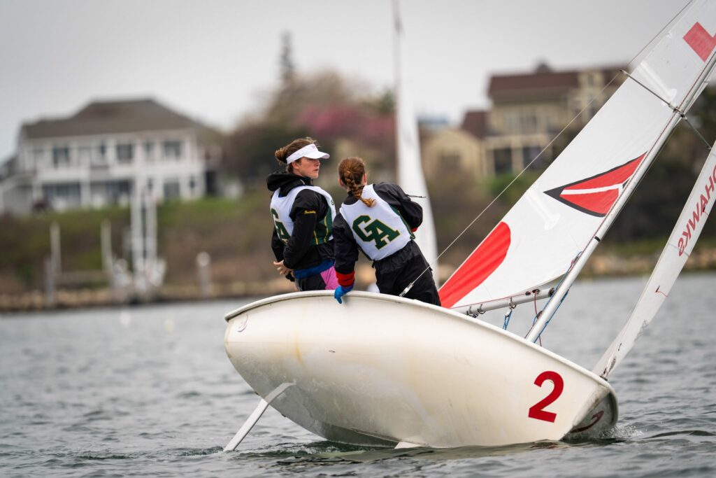 Two people wearing numbered vests sail a small white sailboat numbered “2” on the water, leaning to balance as the boat tilts. Houses and trees appear in the blurry background.
