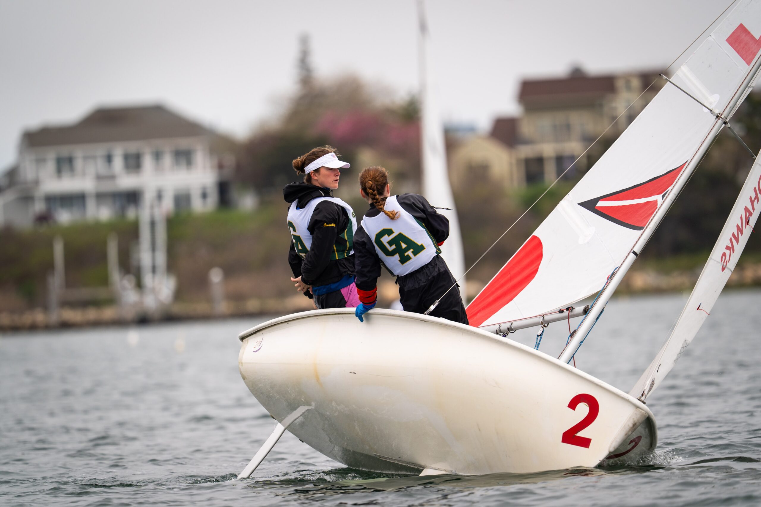 Two people wearing numbered vests sail a small white sailboat numbered “2” on the water, leaning to balance as the boat tilts. Houses and trees appear in the blurry background.