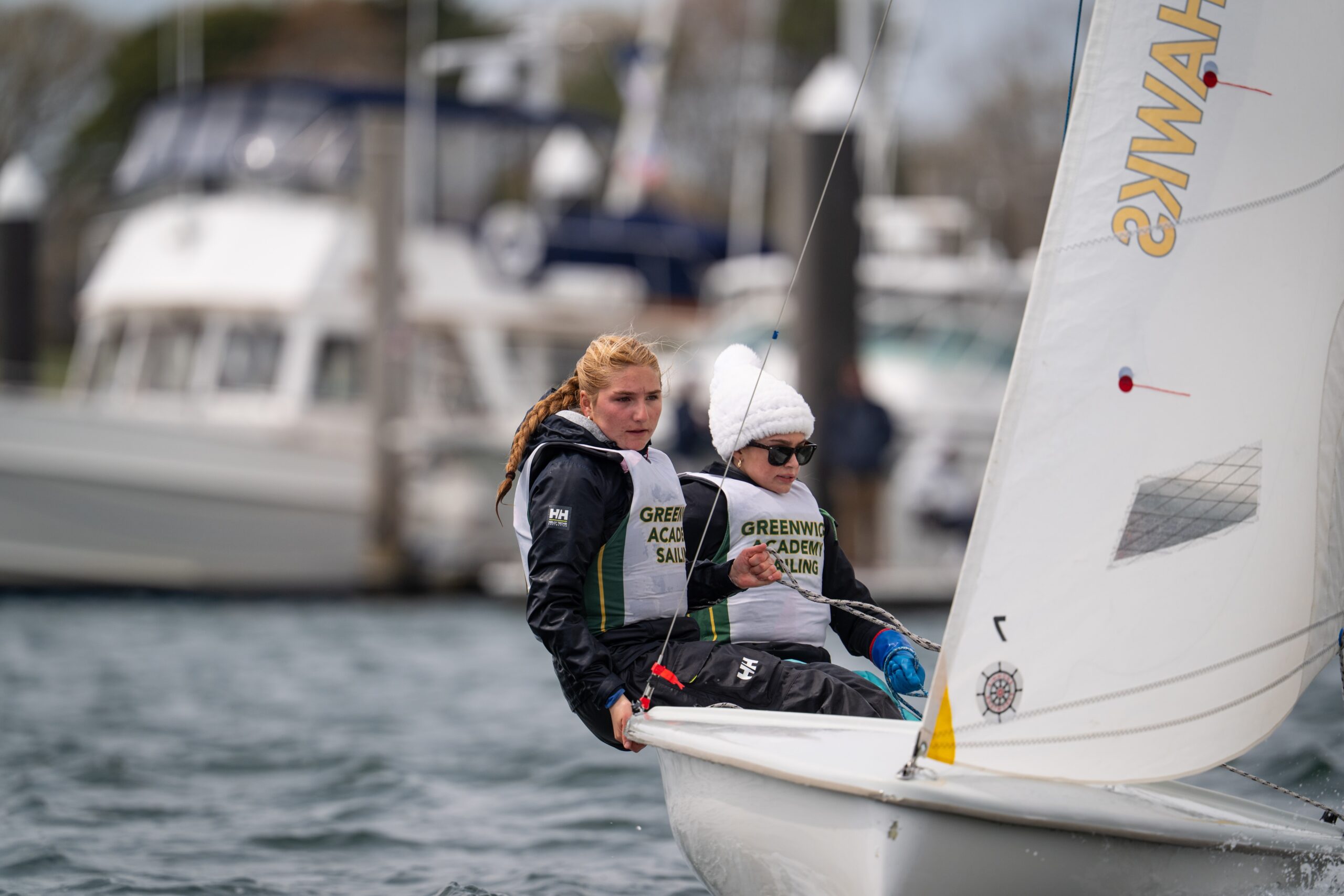 Two young sailors wearing "Greenwich Academy Sailing" vests steer a small sailboat on choppy water, with boats and a marina in the background. One sailor has red hair and the other wears a white hat and sunglasses.