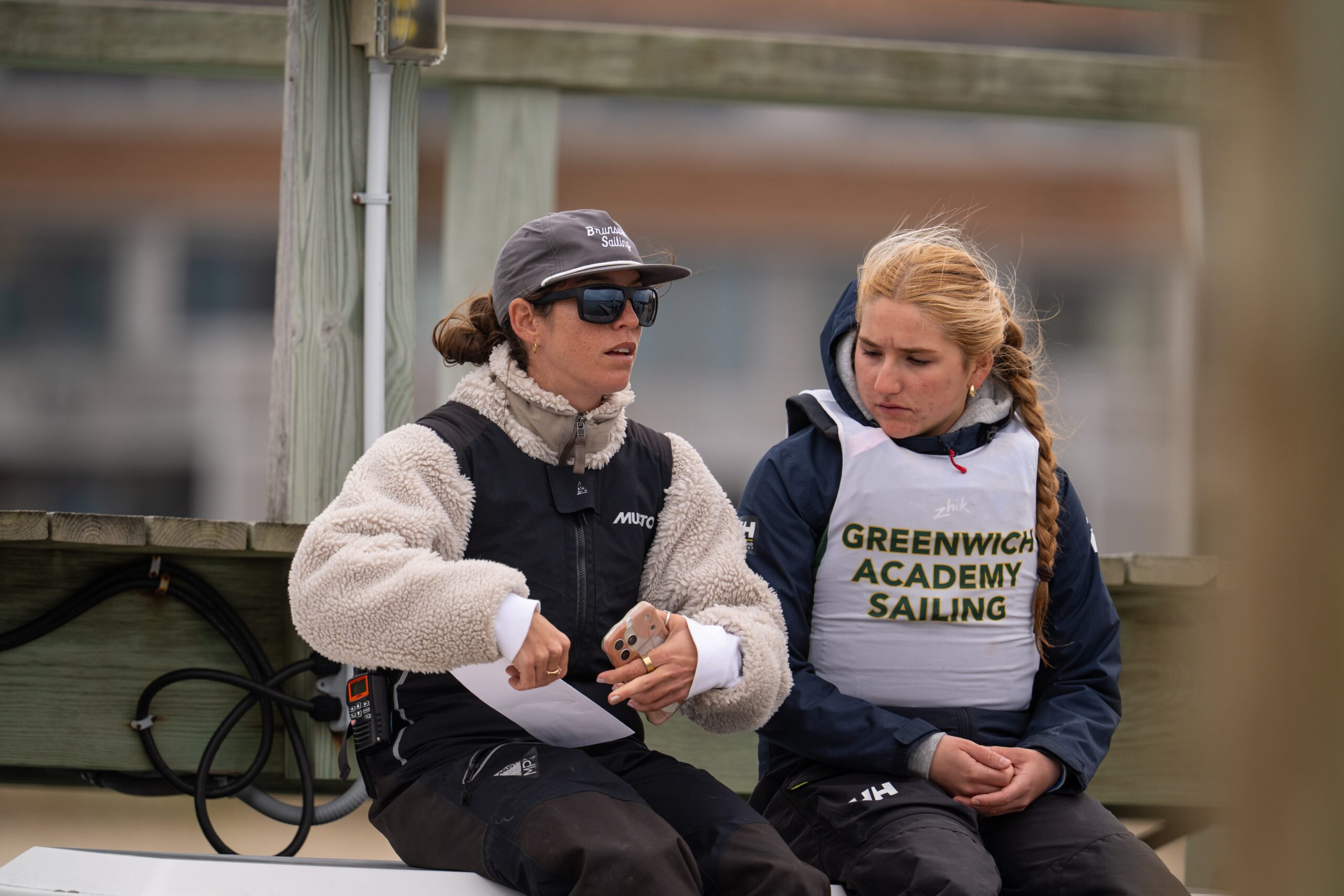 Two people in sailing gear sit on a boat. One gestures while holding a phone and paper, the other looks at them. The second person's vest reads “Greenwich Academy Sailing.” Both appear focused and serious.