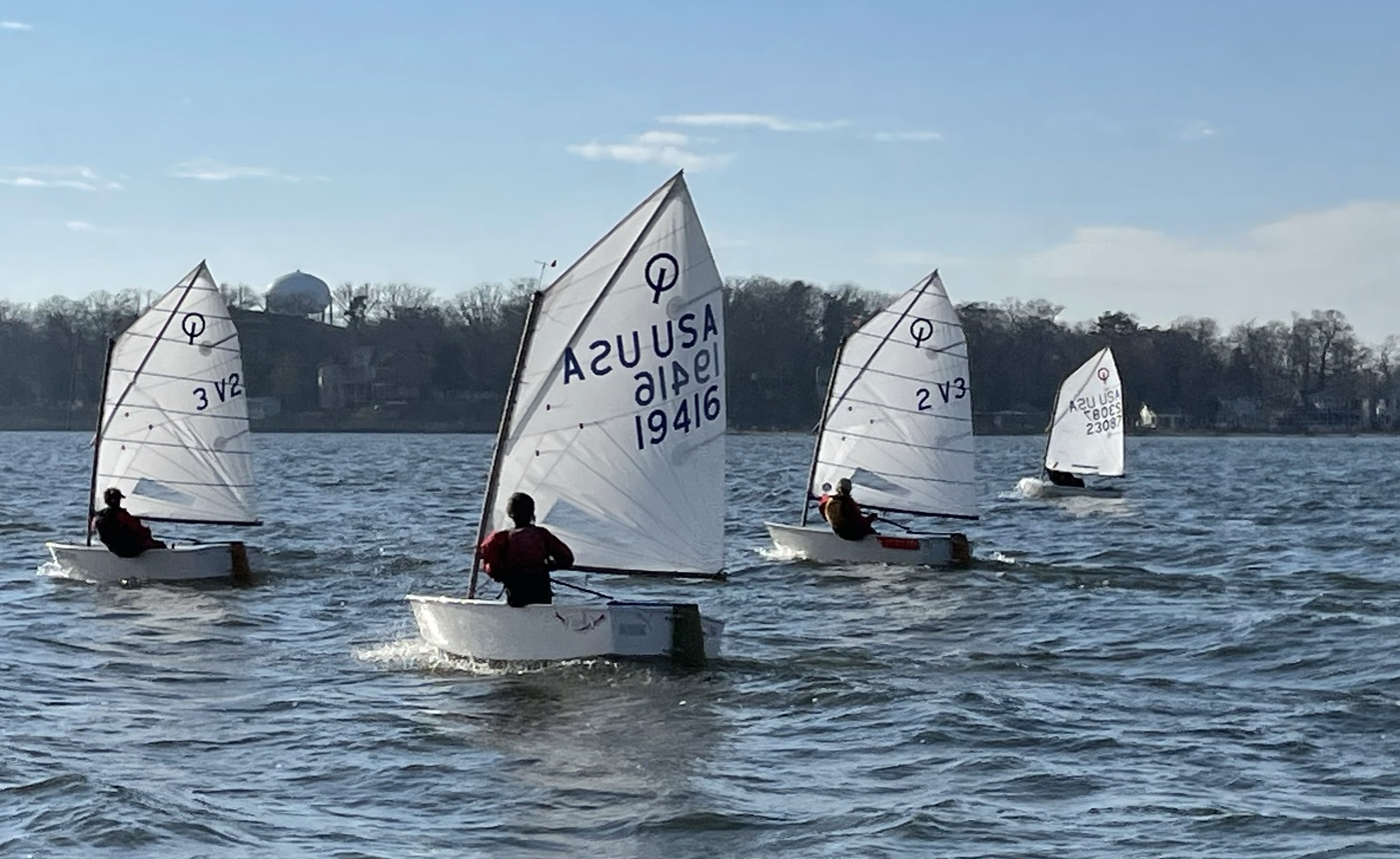 Four small sailboats with white sails and young sailors move across a lake, with trees and a building visible in the background under a clear sky.