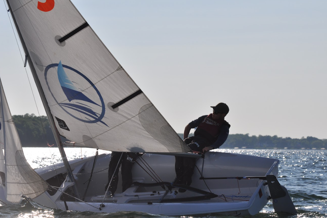 A person leans out of a small sailboat, holding the mainsheet as the boat tilts on a sunny lake with trees in the background.