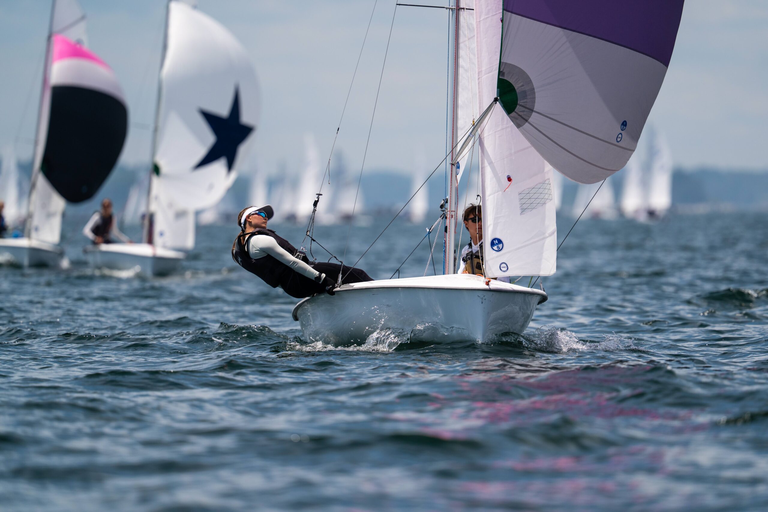 Two people sail a small boat on choppy water, leaning back to balance. Other sailboats with colorful sails are visible in the background under a cloudy sky.