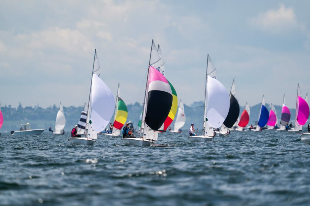 Several sailboats with colorful sails race on a slightly choppy body of water under a cloudy sky, with a distant shoreline visible in the background.