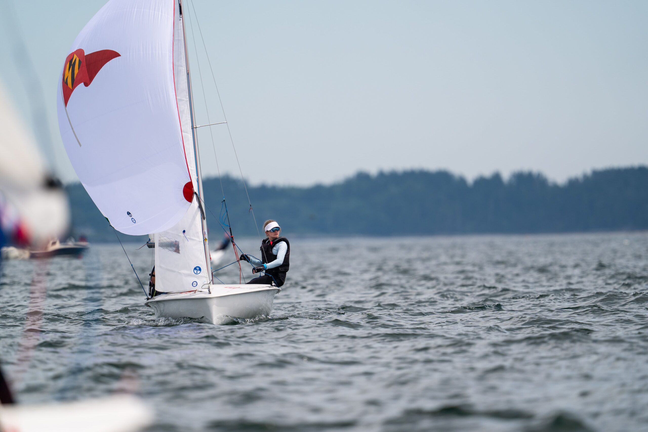 A person in sailing gear navigates a small sailboat with a white spinnaker on open water, with a forested shoreline in the background and other boats partially visible nearby.