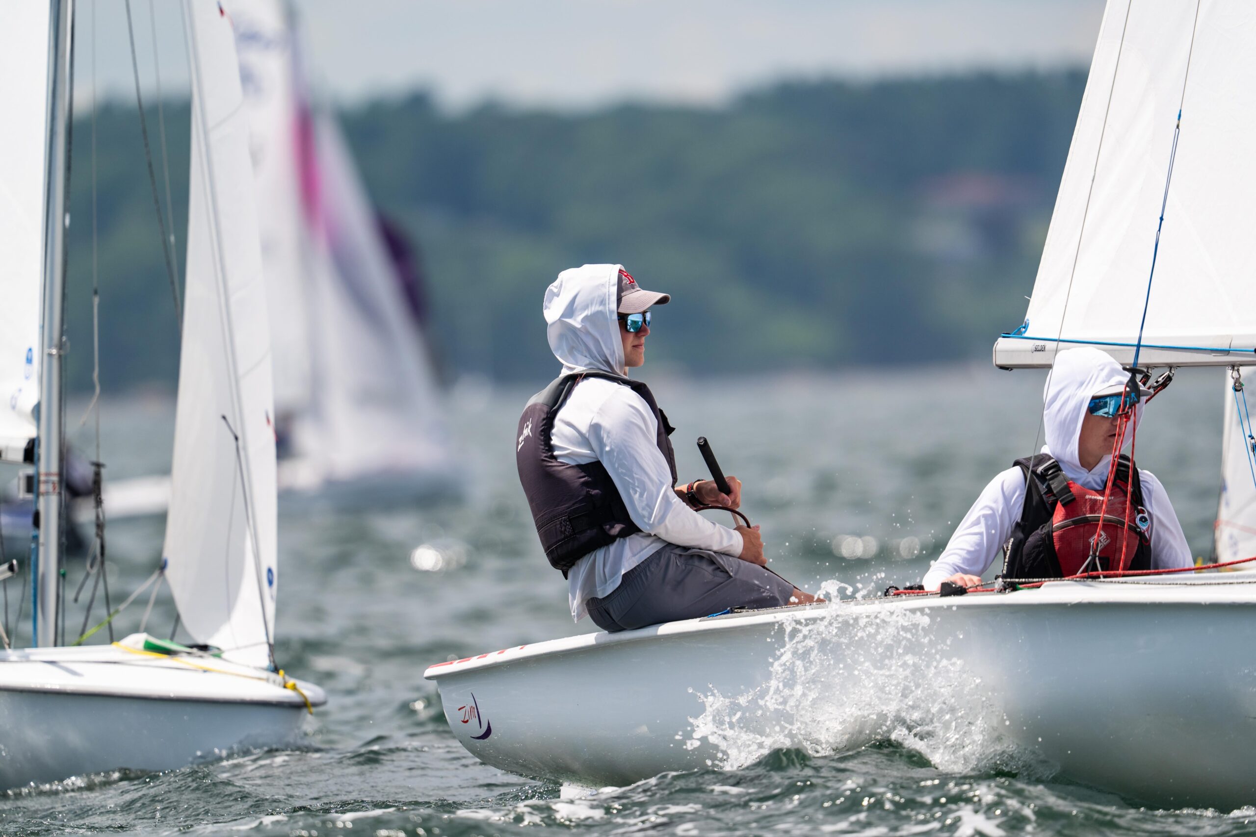 Two people in sun-protective hooded clothing sail a small white boat on choppy water during a race; other sailboats and a blurred shoreline are visible in the background.