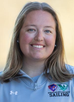 A woman with straight, shoulder-length hair smiles at the camera, wearing a light gray quarter-zip jacket with 'Sailing' and college logos on the chest. The background is softly blurred.
