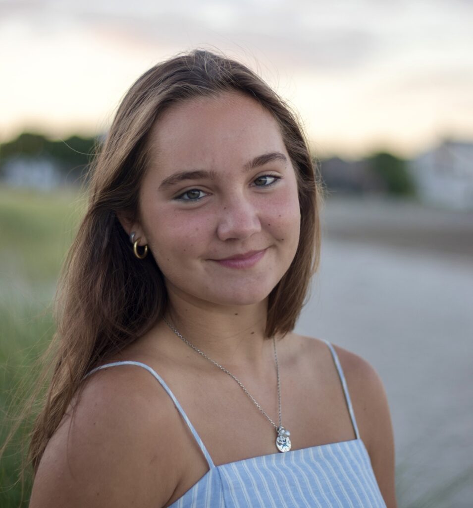 A young woman with long brown hair and blue eyes smiles slightly, wearing a blue-and-white striped dress and a necklace, standing outdoors near a beach at sunset.