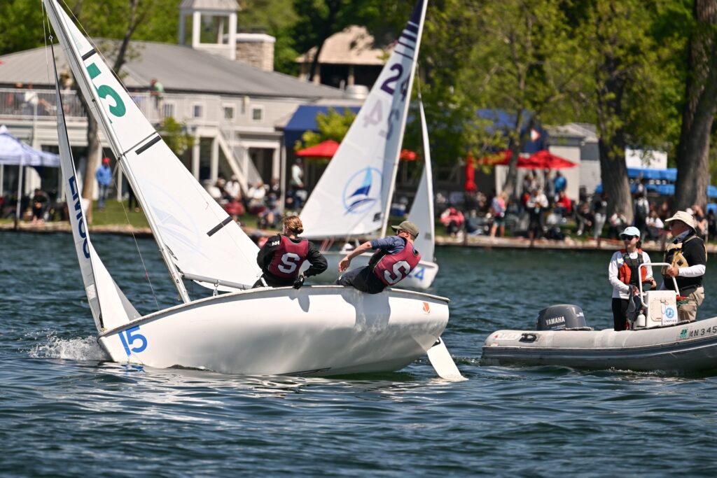 Two sailboats with teams wearing life vests compete on a lake near a shoreline, while people in a motorboat observe. A crowd and a building are visible in the background.