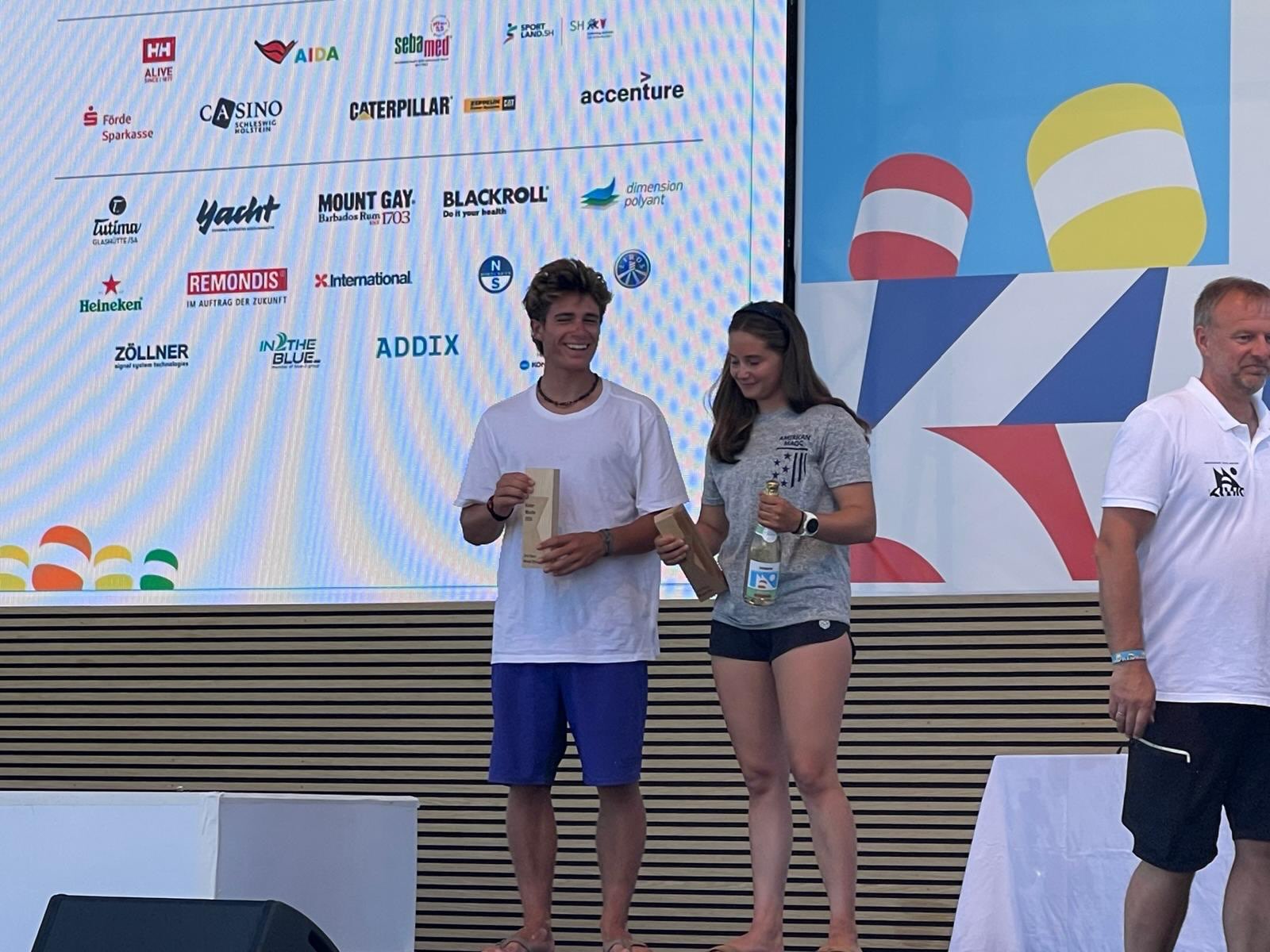 Two young people, a man and a woman, stand barefoot onstage holding brown envelopes and smiling. Behind them is a large sponsor board and a man in a white polo shirt standing to the right.