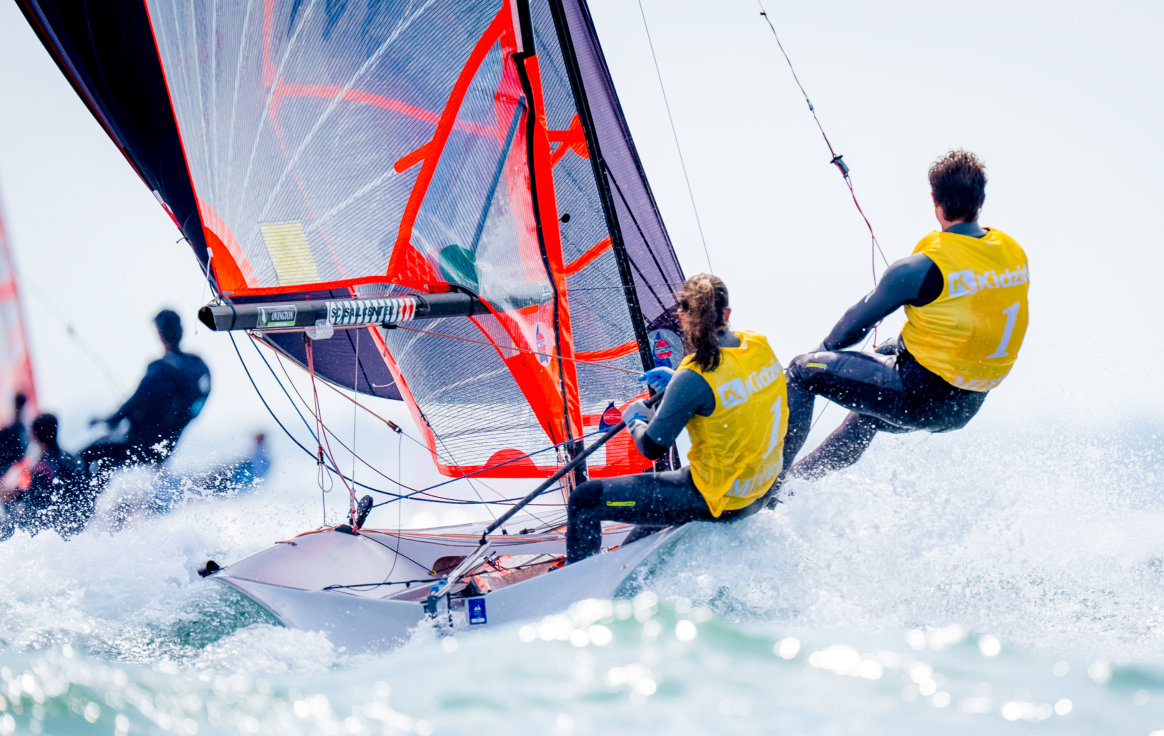 Two sailors in yellow vests race a small sailboat, leaning out over the water. The bright sail catches wind, with waves splashing around the hull. Other boats are visible in the blurry background.