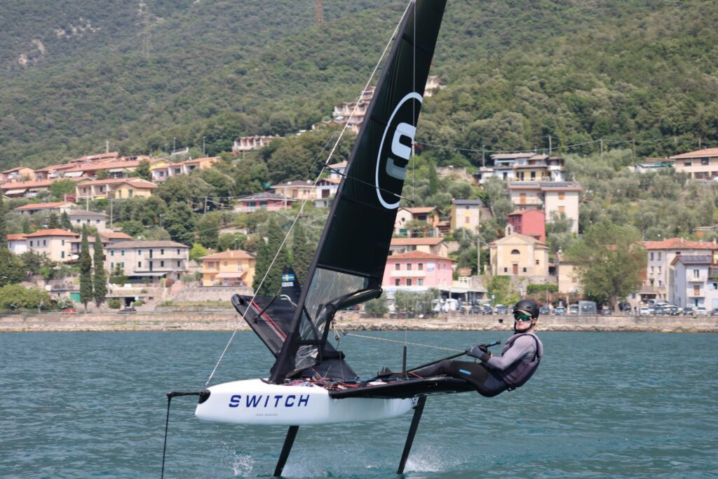 A person sails a small, sleek racing boat labeled 'SWITCH' on blue water, with green hills and colorful houses in the background. The boat has a black sail and appears to be lifted above the water on hydrofoils.