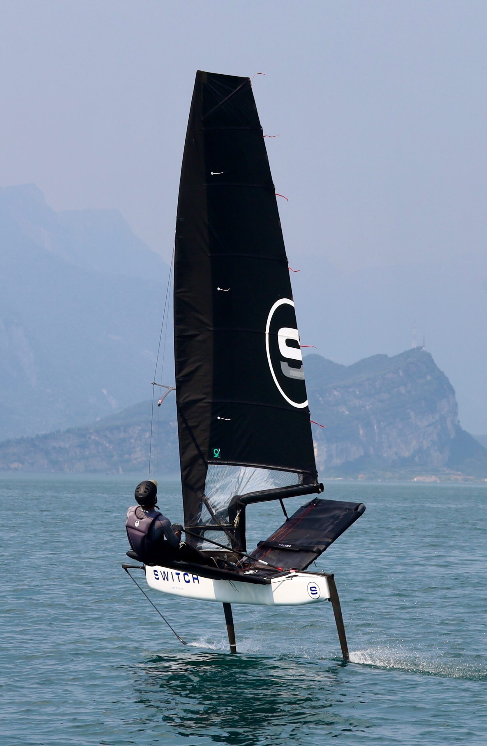 A person sails a sleek black hydrofoil sailboat labeled "SWITCH" over calm blue water, with distant mountains and a hazy sky in the background.