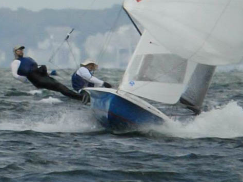 Two sailors in blue and white gear steer a small sailboat at an angle on choppy water, with one person leaning out over the side as the boat cuts through waves. Blurred shoreline is visible in the background.