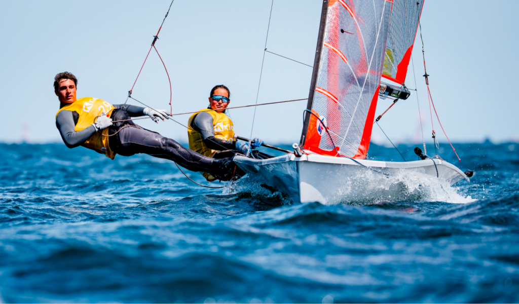 Two sailors in yellow vests lean out from a small sailboat, navigating choppy blue water under a clear sky, with the sails catching the wind.